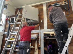 Zwei Monteure installieren die Schaltzentrale einer Bejola-Sprühnebel-Anlage im Vorraum einer Sattelkammer. Die Besonderheit hier: Die Komponenten werden platzsparend im höheren Bereich der Wand installiert, über der Tür zur Sattelkammer