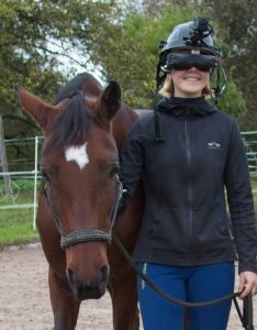 Eine junge Frau führt ein Pferd. Auf dem Kopf trägt sie den Tieraugensimulator, bestehend aus Helm und Visier